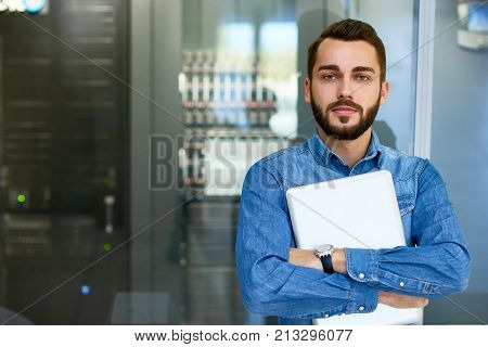 Portrait of beraded systems administrator posing holding laptop and looking at camera standing against server cabinet, copy space