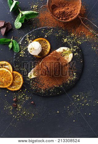 Exquisite french dessert. Chocolate fondant with creme anglaise and vanilla ice cream on round slate with pistachio crumble. Black background with cocoa, mint and sieve, top view, copy space