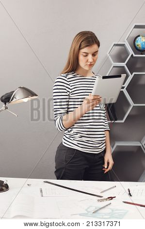 Vertical portrait of young profeciaonnal good-looking architech girl with brown hair in striped shirt and black jeans standing near table looking in digital table, looking through costumer of commision