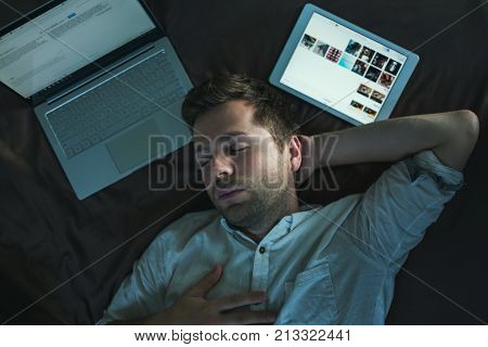 Tired caucasian young man in white shirt sleeping and keeping one hand above head, lying on bed near laptop. He is tired of work problems.