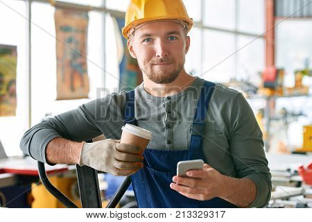Portrait of young bearded worker taking break in factory workshop looking at camera  while using smartphone holding coffee cup