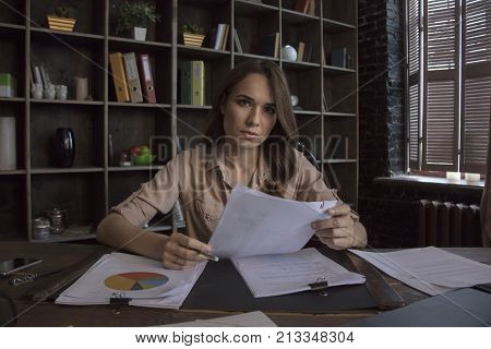 Conducted calculations. Young businesswoman sitting in studio with wistful face behind wooden table with documents with charts and looking forward. Creative startup in studio