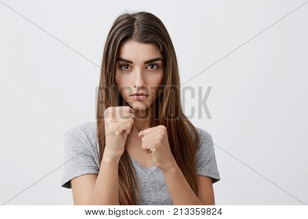Portrait of young serious attractive caucasian female with dark long hair in stylish gray t-shirt holding hands i fight pose, looking in camera with mean and concentrated expression, ready to punch you in face