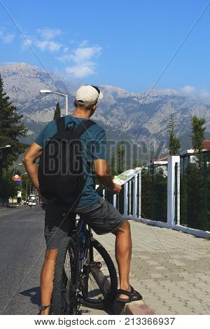 a man stands on the road in the village and looks at the tourist map dressed in a T-shirt and cap. a man with a backpack stands on a background of high mountains.