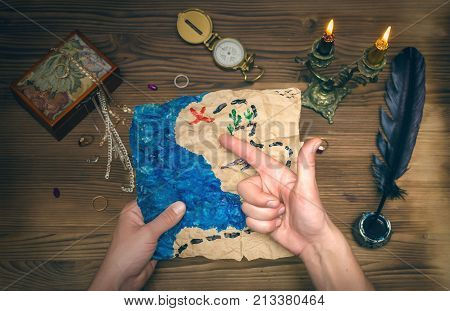 Crumpled treasure map and pirate hat on wooden table. Treasure hunt. Pirate man showing the way to the gold chest marked on the treasure map with his hand.