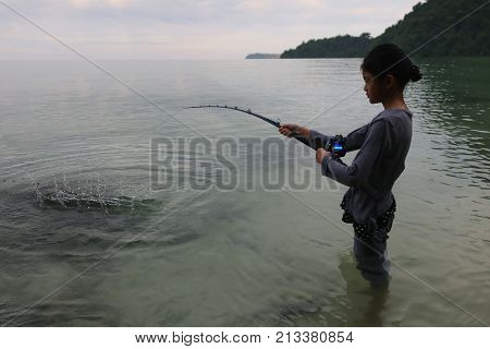 Asian girl standing and fishing with rod and reel in the sea near the beach with red sky sunset background