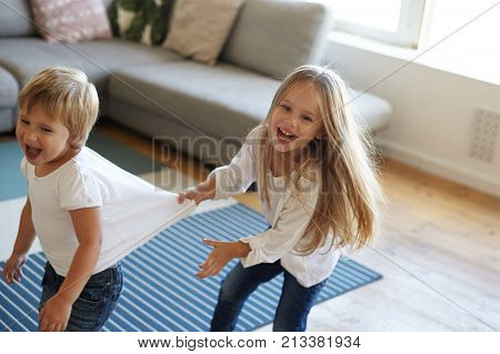 Happy overjoyed female child laughing and screaming with joy pulling her excited little sister by white t-shirt while playing tag at home or kindergarten feeling joyful. Kids leisure and fun
