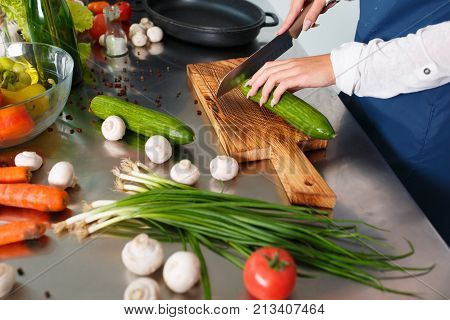Close-up of a girl's hand slice a cucumber on a cutting board. On the kitchen table scattered vegetables are champignons, green onions, pepper peas, tomato.