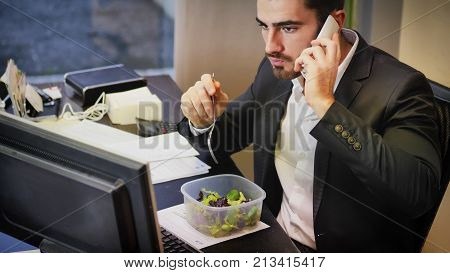 Elegant man in suit having lunch at table in office while talking on phone and watching computer.