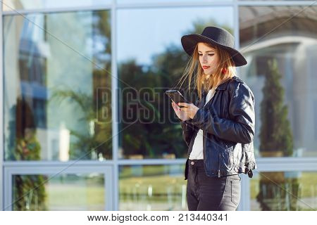 Stylish hipster girl wears black leather jacket and holding phone before modern building