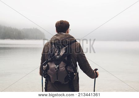 Back view of man with backpack and trekking sticks exploring landscape with misty lake in mountains.