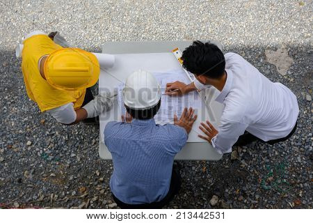 Top view of young engineer present constrcuted paper blueprint plans to site manager and foreman at construction site. Business Engineering Concept for team work of heavy industry.