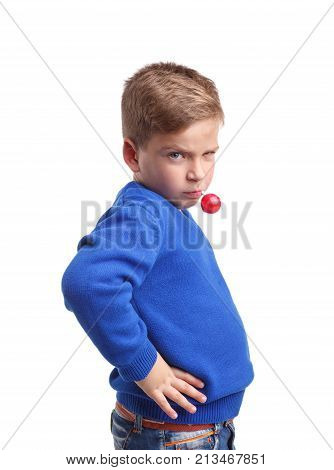 A boy in a blue sweater with a suspicious glance is holding a stick with a lolli-pop in his mouth. Standing sideways on a white isolated background