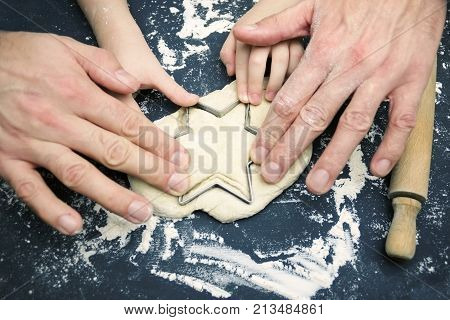 A photo from above of father and child hands cutting the star with a cookie cutter on the wooden table. An overhead photo of father's and kid's hands flour wheat dough cookie cutter rolling pin