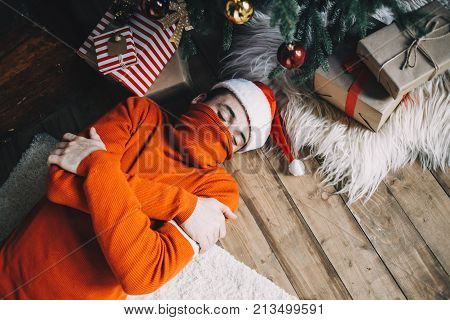 Guy is lying on the floor near christmas tree with his arms crossed and sleeping. He has a hat on his head and a sweter's neck on his mouth. It's apparently cold in room after in the morning after celebrating Christmas eve. Cut view