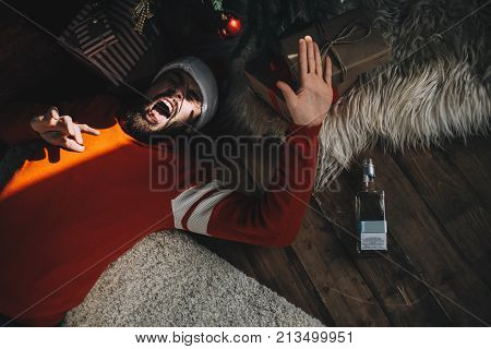 One adult is lying on the floor near pine tree in room in the sunny morning. He is trying to cover his face from the sunlight. Plus this guy has a morning hangover after drinking too much alcohol last evening during the Christmas celebration. Cut view