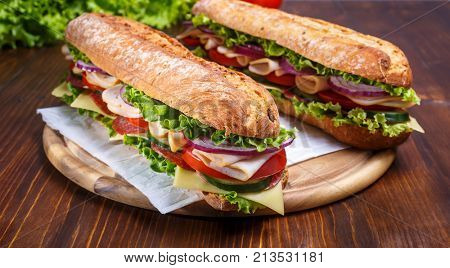 Two baguette sandwiches with salami, turkey breast, cheese, lettuce, tomatoes and onion on a cutting board