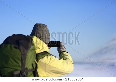 Back view of unrecognizable male in hat and cout with fur hood carrying stylish backpack taking picture of amazing winter landscape using copyspace mobile phone while walking alone on sunny day.