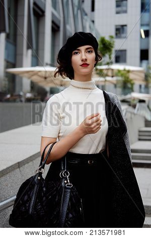 Stylish young female office worker in classic outfit in the city center wearing white turtle neck black beret and cardigan. Business city coffee break.