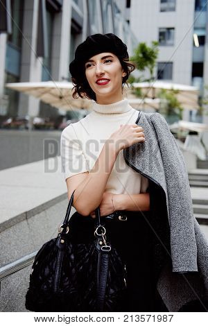 Stylish young female office worker in classic outfit in the city center wearing white turtle neck black beret and cardigan. Business city coffee break.