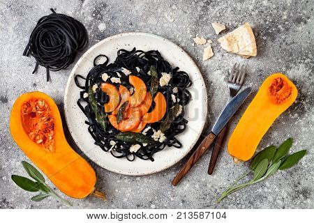 Black pasta with roasted butternut squash parmesan cheese and fried sage. Concrete background top view flat lay. Halloween black and orange party dinner concept
