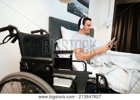 The invalid lies in bed and looks something on the tablet. He smiles. because he is happy. Next to his bed is his black wheelchair. It lies on a large white step.