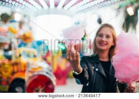 Soft focus shot of young and happy hipster millennial girl or teenager standing next to attraction ride at festival or carnival fair surrounded by colors and lights holds pink cotton candy treat smiles