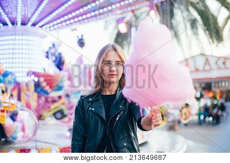 Happy attractive and beautiful young woman or female teenager with blonde hair wears black leather jacket stands in front of attraction ride at festival or carnival and holds huge pink cotton candy