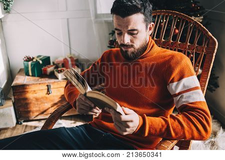 Good-looking and handsome guy is sitting in the rocking chair beside a Christmas tree and reading an old book. He wears red Christmas poleneck sweater. He is enjoying time spending like that. Close up. Cut view