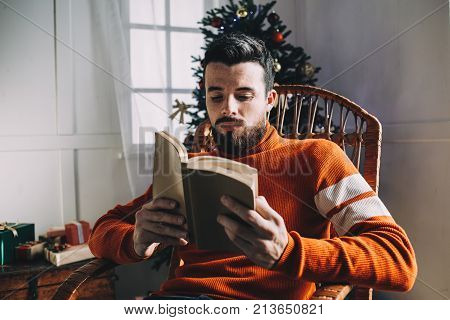 Another picture of good-looking guy sitting in the rocking chair at the Christmas tree and reading interesting book. He has almost finished reading it. Man decided to read a book while he is waiting for his friends to come to celebrate Christmas.