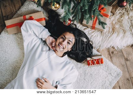 Incredible and stunning young woman is lying on the white carpet between Christmas gifts and beautiful green pine tree. She is relaxing and enjoying her time spanding like that. Close up. Cut view