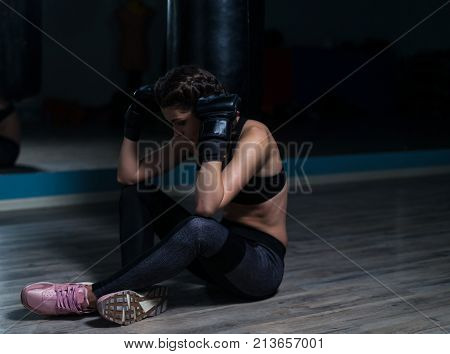 Upset young fighter boxer girl wearing boxing gloves siting on the floor after loosing fight.  She is thinking