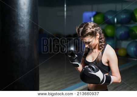 Upset young fighter boxer girl wearing boxing gloves in gym beating punching bag. She is sad