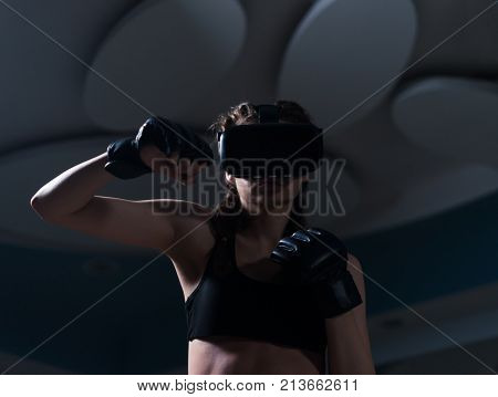 Young fighter boxer fit girl in VR glasses wearing boxing gloves in training. Boxing game in virtual reality 360 degrees. Futuristic gaming. She is punching an enemy