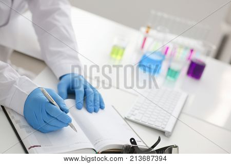 Employee of the chemical laboratory teacher chemist holds a silvery pen in his hand makes notes in the diary records test data from reactions examining test tubes with the substance arm in gloves.