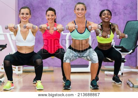 Beautiful athletic women doing squats while holding dumbbells with straight arms forward during functional training group class in a modern fitness club for ladies only
