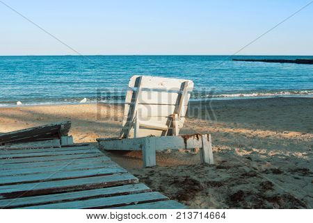One lonely old white vintage wooden sun lounger standing at the abandoned beach after the end of a summen season in the evening sunlight.