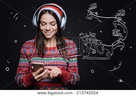 Enjoying jingle bells song. Cheerful amused young woman standing against imaginary drawing on the black wall and expressing happiness while listening to the Christmas songs