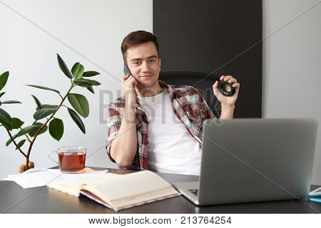 portrait in natural light of a young man in casual clothes sitting on a chair behind a Desk with a laptop while talking on the phone from compressing the expander.