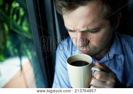 Close up portrait of caucasian man drinking coffee, looking trough window and holding cup. He is sad and upset