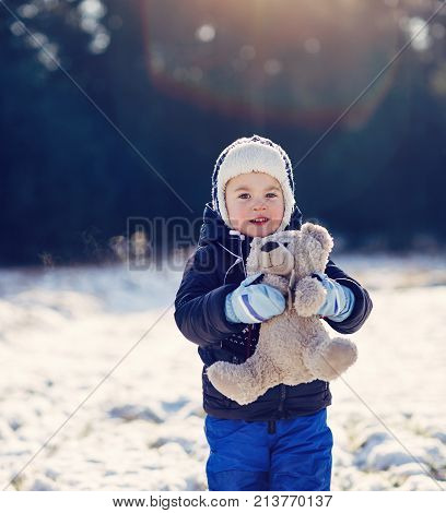 Adorable little boy holding his teddy bear outdoors on a snowy winter's day in foreat with flare