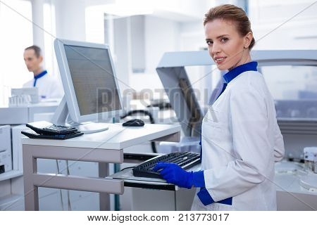 Working atmosphere. Resolve pleasant female scientist smiling at the camera while standing in the lab and typing