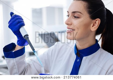 Right dosage. Portrait of friendly smiling laboratory assistant who holding pipette looking at it  and wondering