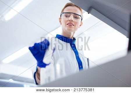 This one. Beautiful serious female laboratory assistant wearing medical glasses and looking down while  taking container from the stand