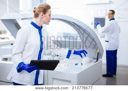 I get it. Good looking curious female laboratory assistant  wanting to take glassware while holding a tablet and posing on the blurred background