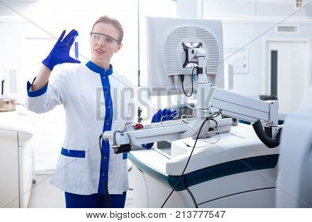 Transferring results. Attractive attentive female lab assistant  standing near advanced medical apparatus while holding test tube and typing
