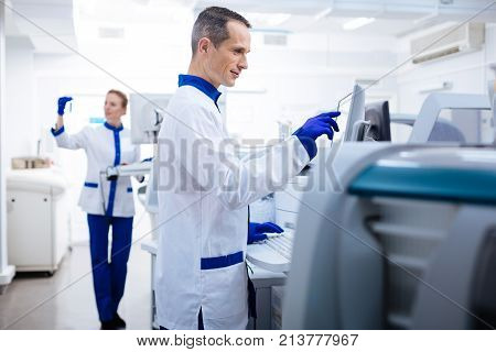 Everything in order.  Positive thoughtful male laboratory assistant  typing and pointing at the screen while posing on the blurred background
