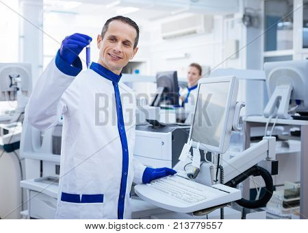 Successful experiment. Happy pleased male researcher achieving necessary reaction while placing his hand on the keyboard and brining up  glassware while standing in the lab