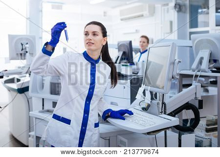 New achievement. Smart merry female scientist putting a hand above the keyboard while studying new sediment which forming in the glassware