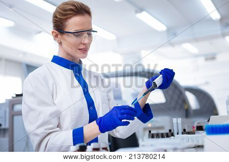Caution with chemicals. Blond attractive female scientist   looking down and  holding pipette when posing against blurred background